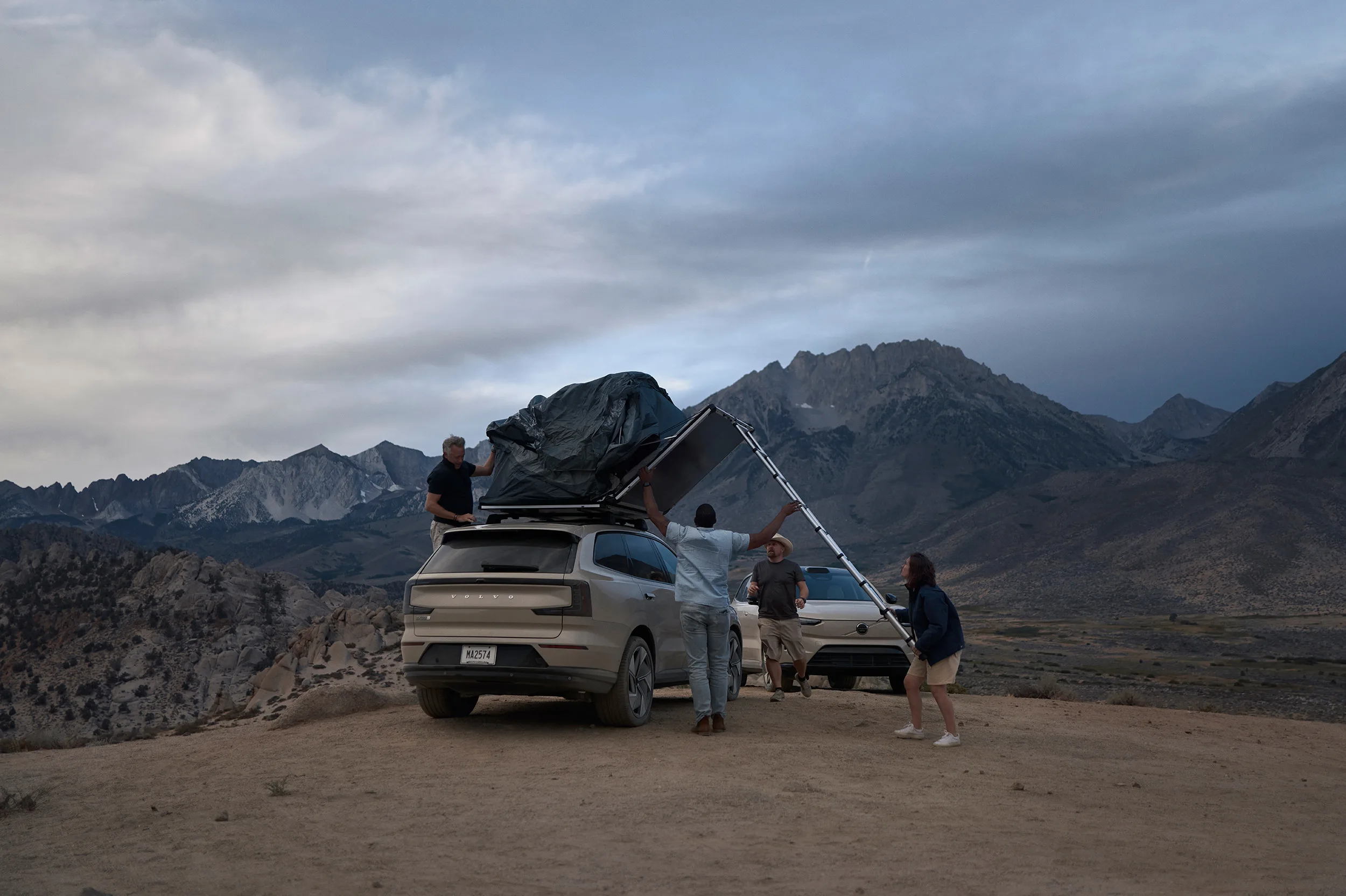 Four people set up a rooftop tent on an SUV parked on a hilltop, with mountains in the background under a cloudy sky. Another car is parked nearby, and the group appears to be preparing for camping.