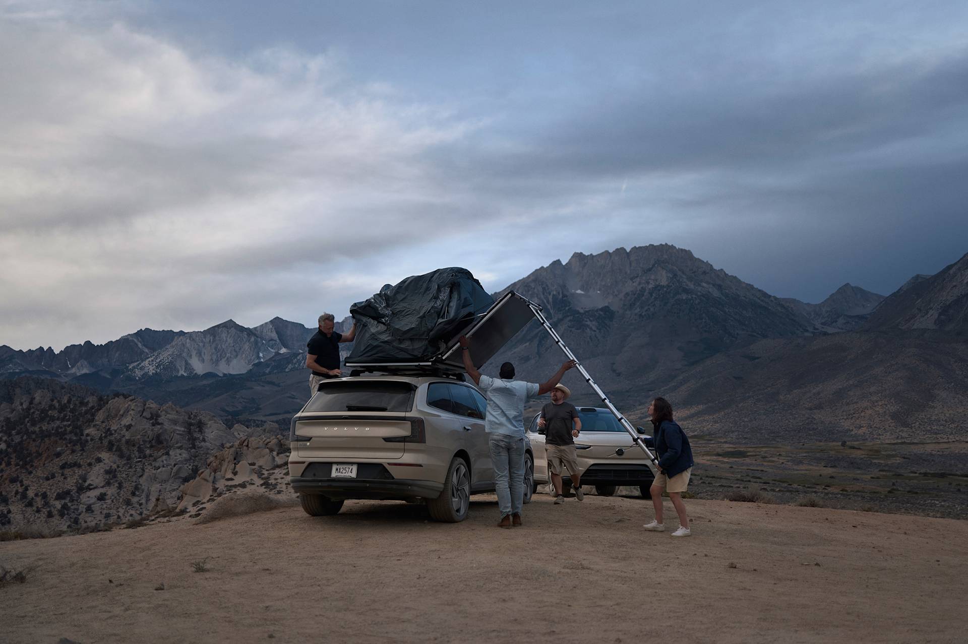 Four people set up a rooftop tent on an SUV parked on a hilltop, with mountains in the background under a cloudy sky. Another car is parked nearby, and the group appears to be preparing for camping.