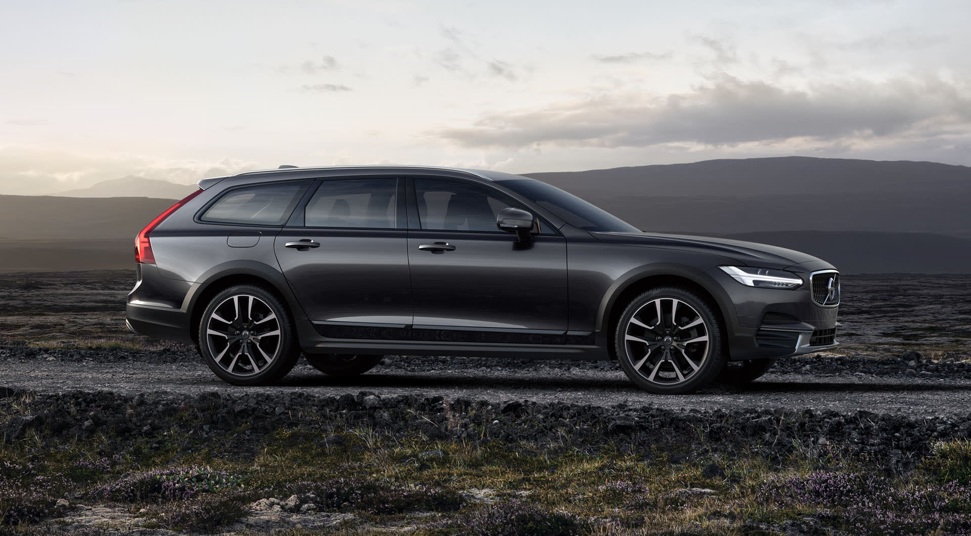A dark gray Volvo station wagon parked on a gravel road in a rugged landscape, with hills and a cloudy sky in the background.