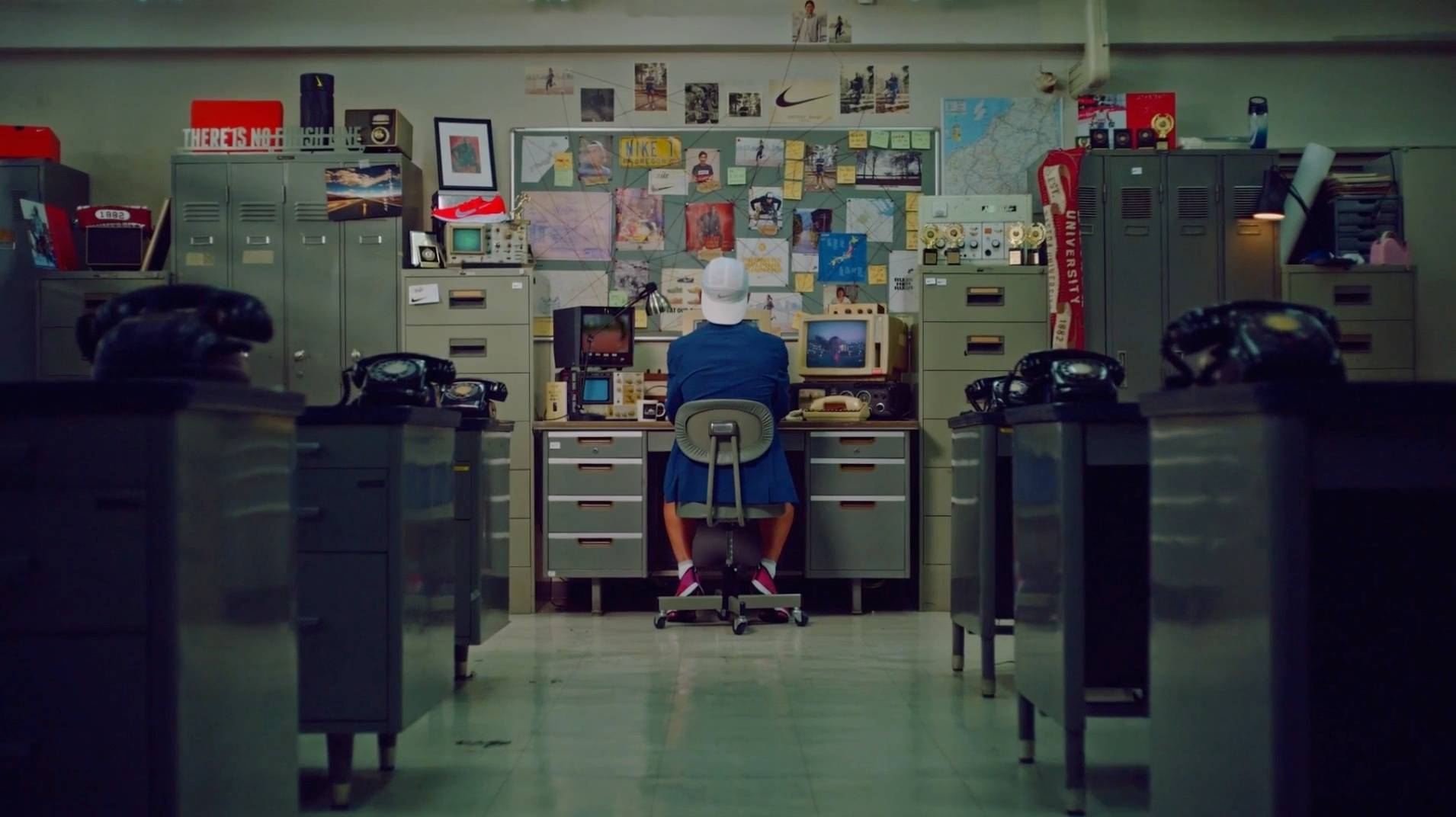 A person sits at a cluttered desk in an office, facing a wall covered with photos, notes, and maps. Filing cabinets and vintage black telephones line both sides of the room. The atmosphere feels retro and organized.