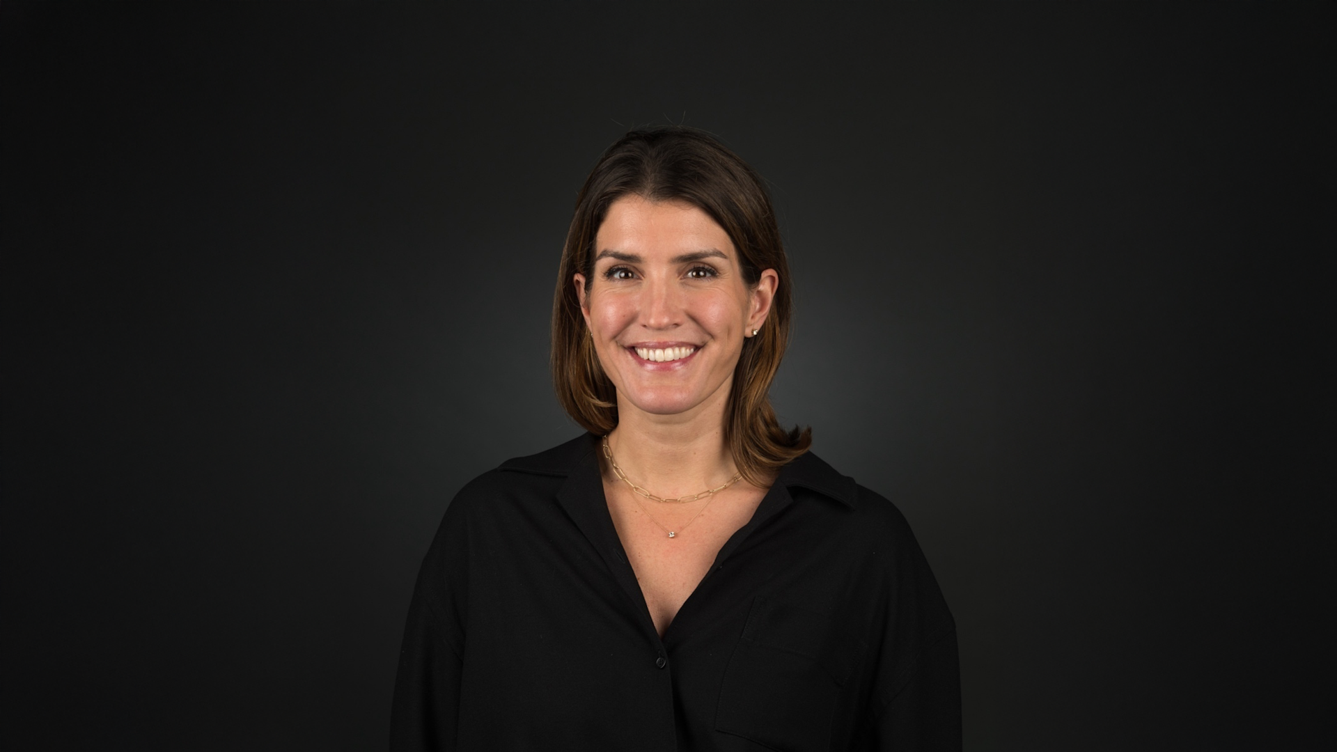 A woman with brown hair wearing a black shirt and a gold necklace stands smiling against a plain dark background.