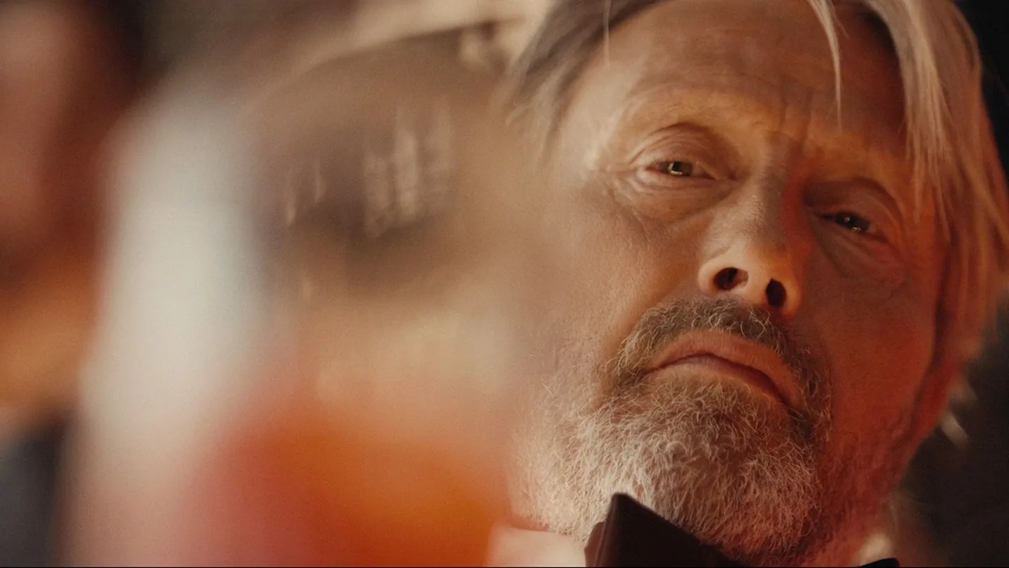 A close-up of a bearded man with gray hair and a serious expression, wearing a black bow tie, gazing past a blurred glass with an amber-colored drink in the foreground. Warm lighting creates a dramatic mood.