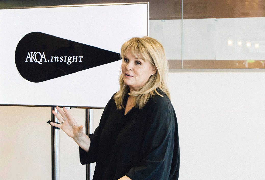 A woman speaks in front of a screen displaying the logo AKQA Insight. She gestures with one hand, wearing a loose black top, with light-colored hair and a neutral expression in a modern, bright room.