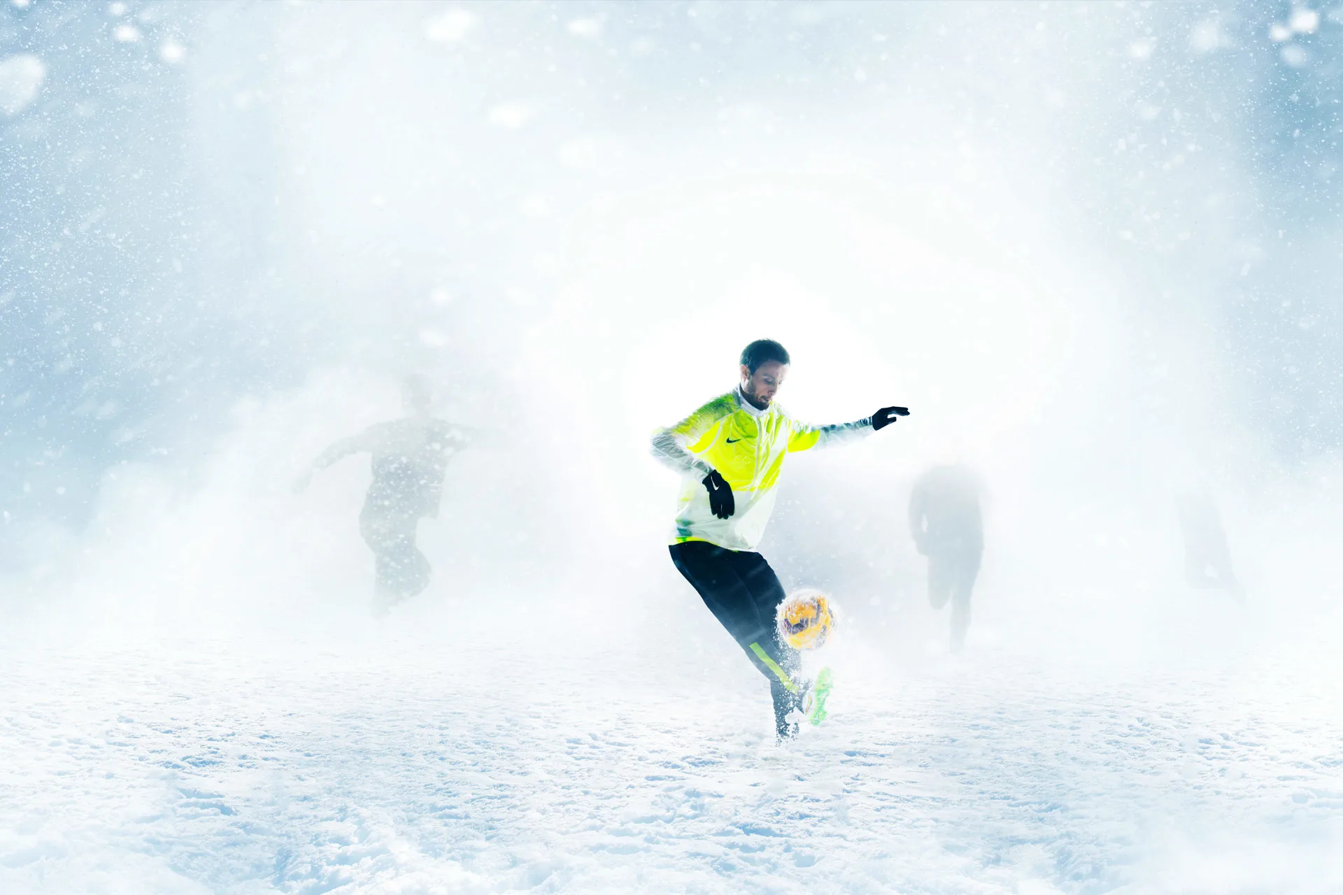 A soccer player in a yellow jersey kicks a ball on a snowy field, with three blurred players in the background and heavy snow falling all around.