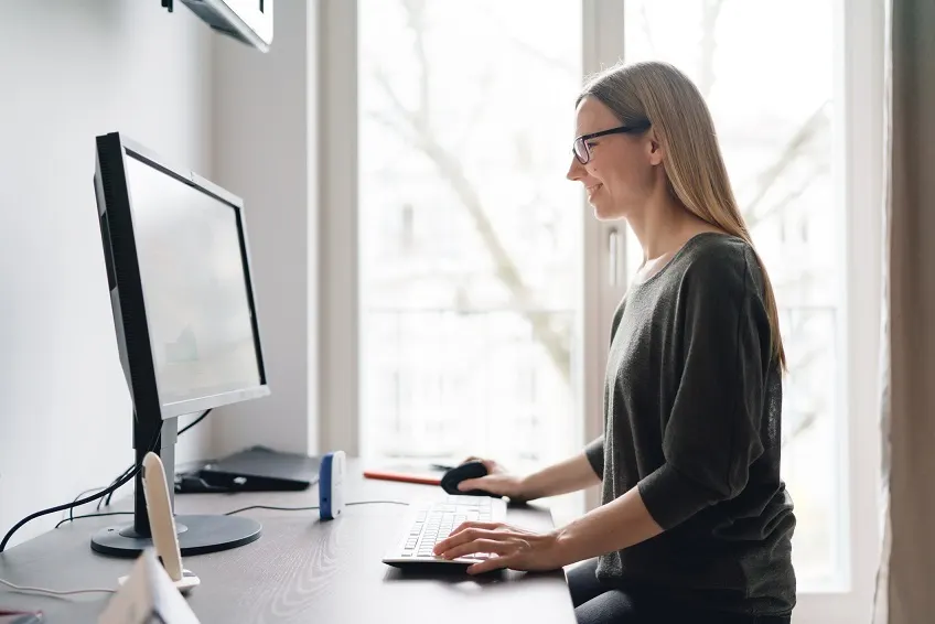 A woman wearing glasses sits at a desk, smiling and using a desktop computer in a bright room with large windows in the background.