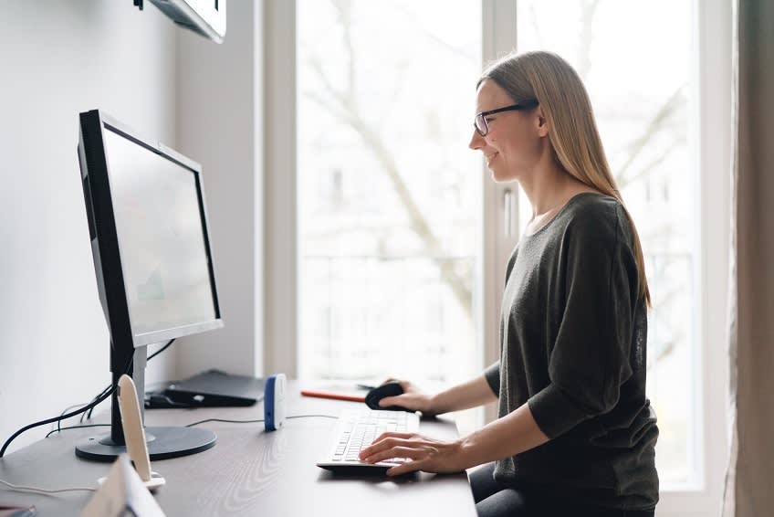 A woman wearing glasses sits at a desk, smiling and using a desktop computer in a bright room with large windows in the background.