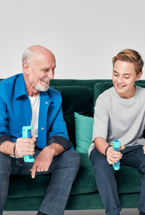 An elderly man and a young boy sit on a green couch, smiling and playing a video game together, each holding a colorful game controller.