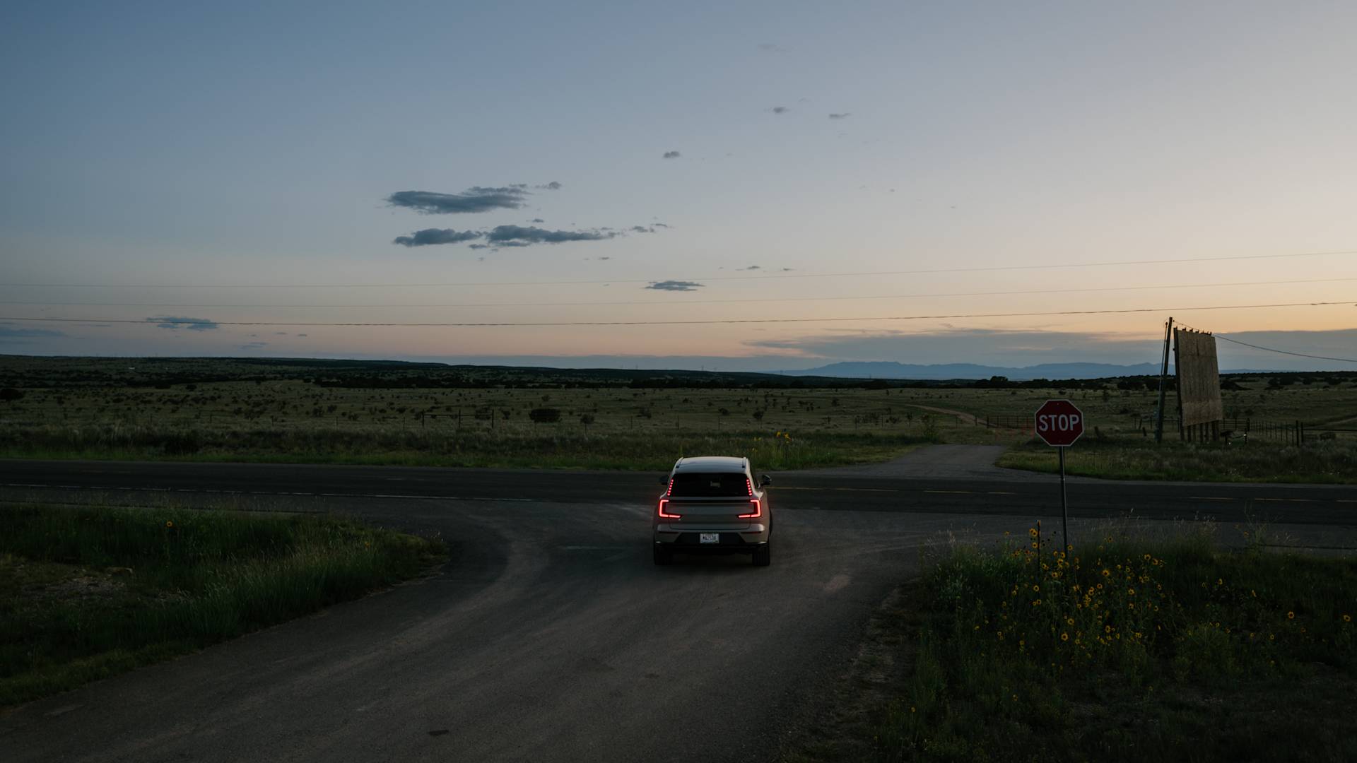 A car with taillights on waits at a stop sign at a rural intersection during dusk, with open fields and a wide sky in the background.