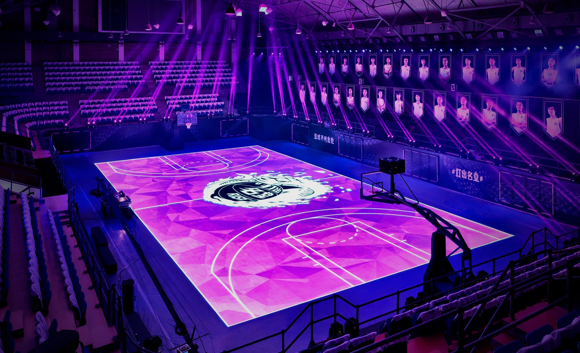 A basketball court illuminated with vibrant purple lights, featuring a digital design on the floor. The stands are mostly empty, and large photos of players are displayed along the wall. The arena has a futuristic atmosphere.