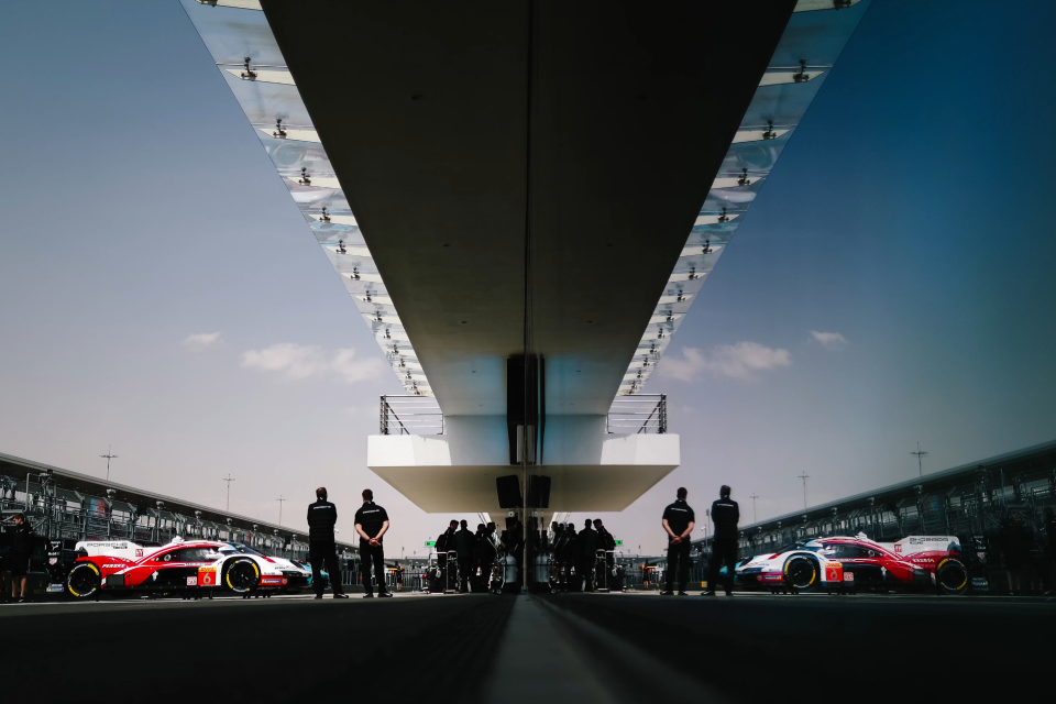 Two race cars and teams prepare under a modern, mirrored canopy at a race track, with people and cars reflected symmetrically on both sides, creating a dramatic, futuristic scene.