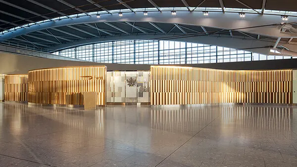 A modern airport lounge entrance with a curved facade featuring vertical wooden slats and warm lighting, set in a spacious area with large windows and a high, arched ceiling.
