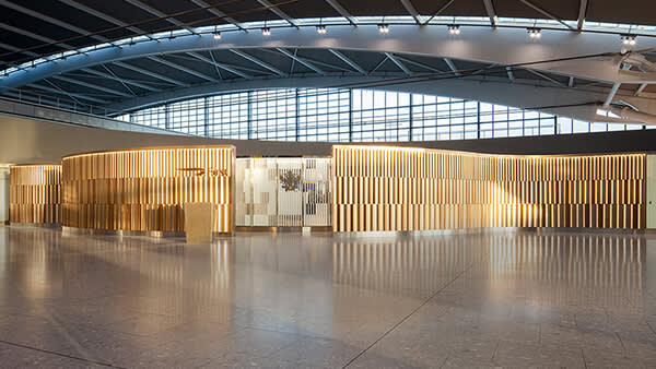 A modern airport lounge entrance with a curved facade featuring vertical wooden slats and warm lighting, set in a spacious area with large windows and a high, arched ceiling.