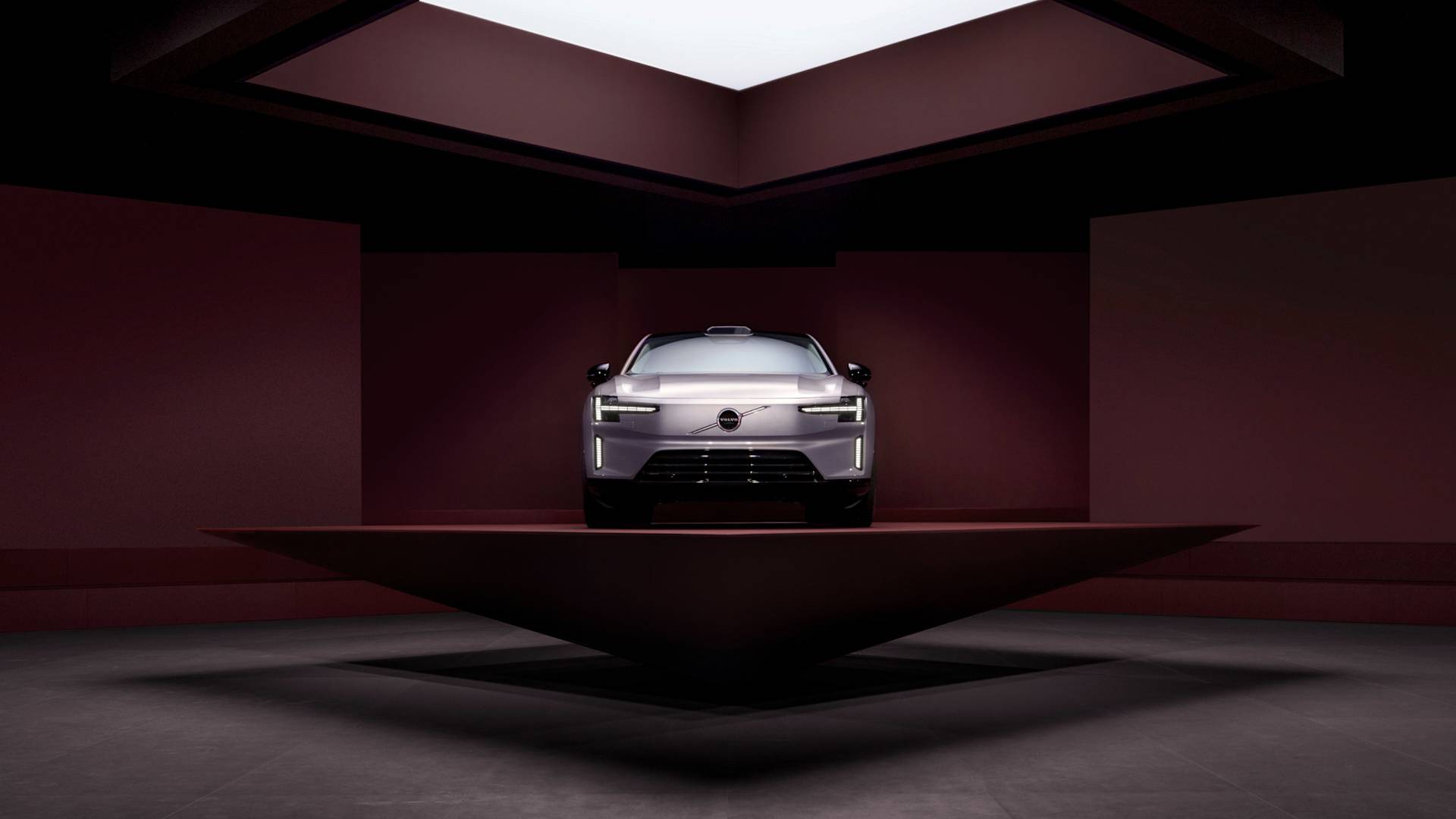 A silver car is displayed on a raised, angular platform in a modern, dark red room with a large square skylight above, highlighting the vehicles front view and sleek design.