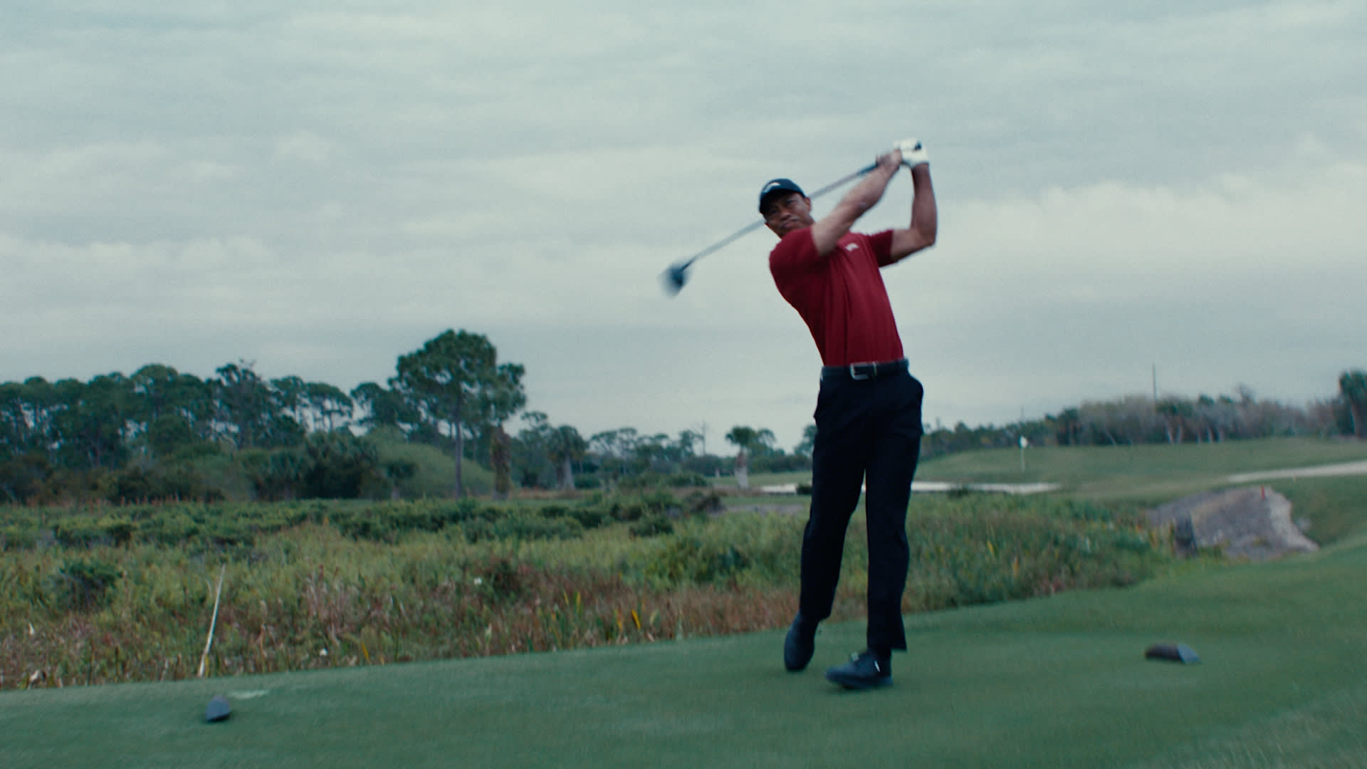 Tiger Woods in a red shirt and black pants finishes a swing on a golf course, surrounded by greenery and trees under a cloudy sky.