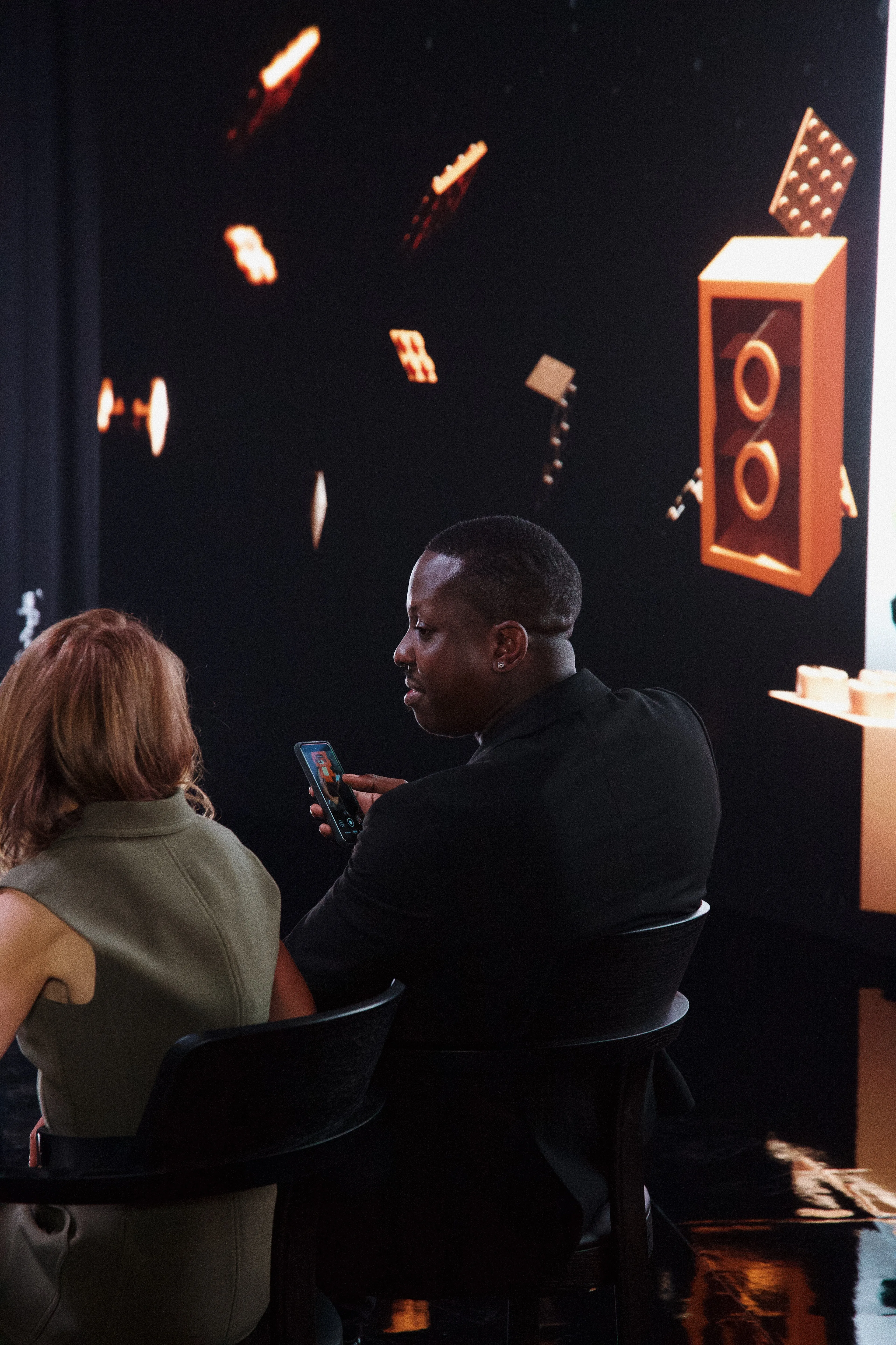 A man in a dark suit and a woman sit side by side, with the man holding a phone. Behind them is a large screen displaying floating 3D objects, including speakers and geometric shapes.