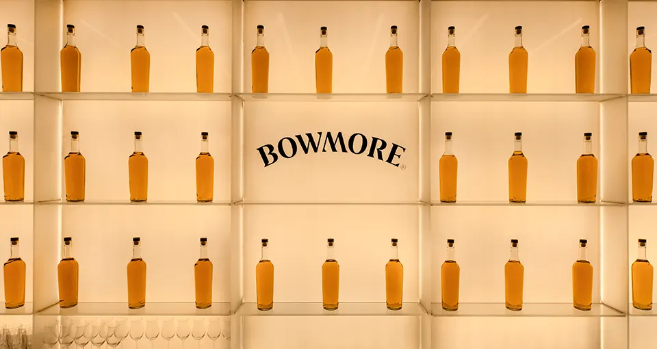 A backlit display of evenly spaced bottles filled with amber liquid, labeled BOWMORE in the center. Empty wine glasses are arranged on the lower shelves.