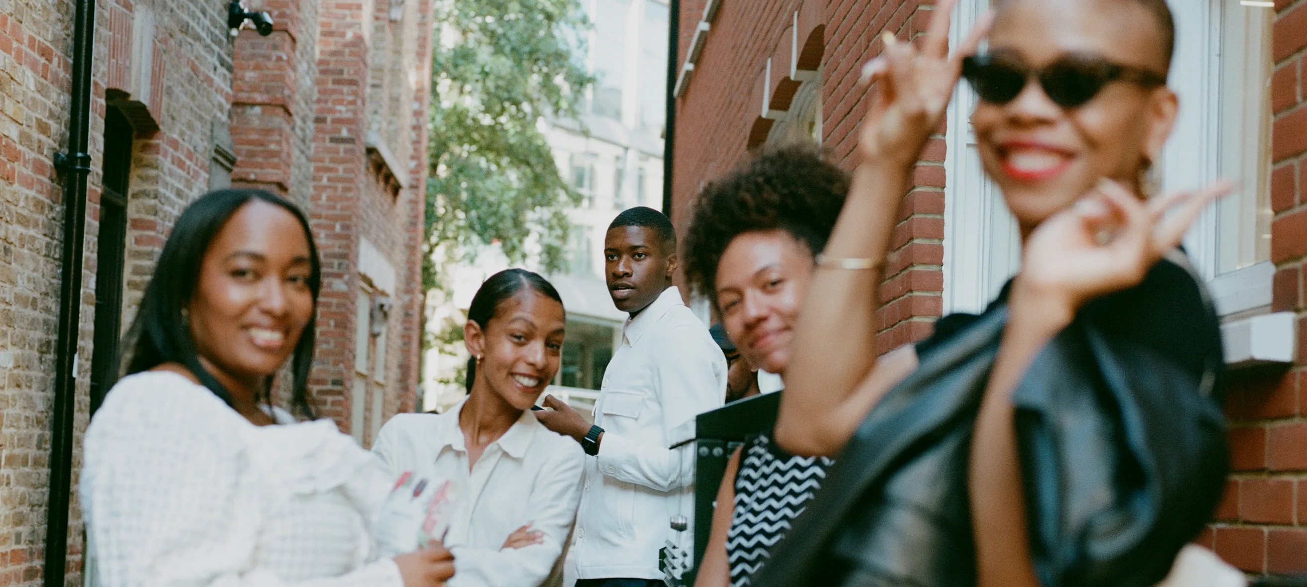 A group of five people stand in a narrow alleyway between brick buildings. Four are smiling and posing for the camera in the foreground, while one person stands in the background looking towards them.