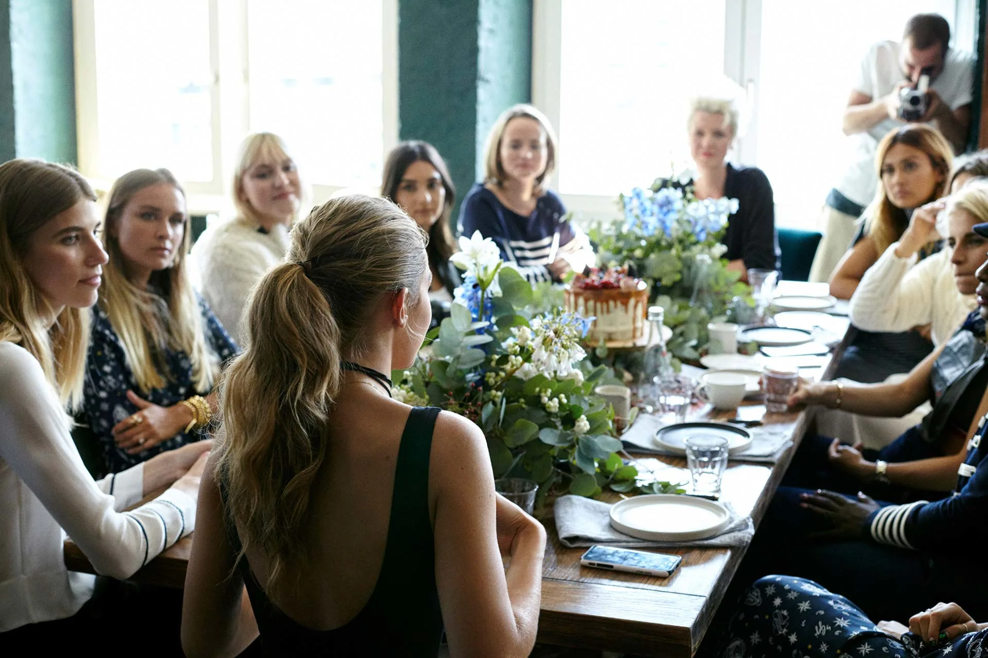 A group of women sit around a wooden table decorated with flowers and food, having a conversation in a bright room while a photographer takes pictures.