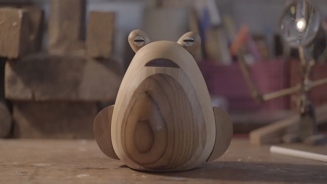 A wooden frog figurine with smooth, rounded shapes sits on a workbench in a workshop. The background includes blurred tools and materials.
