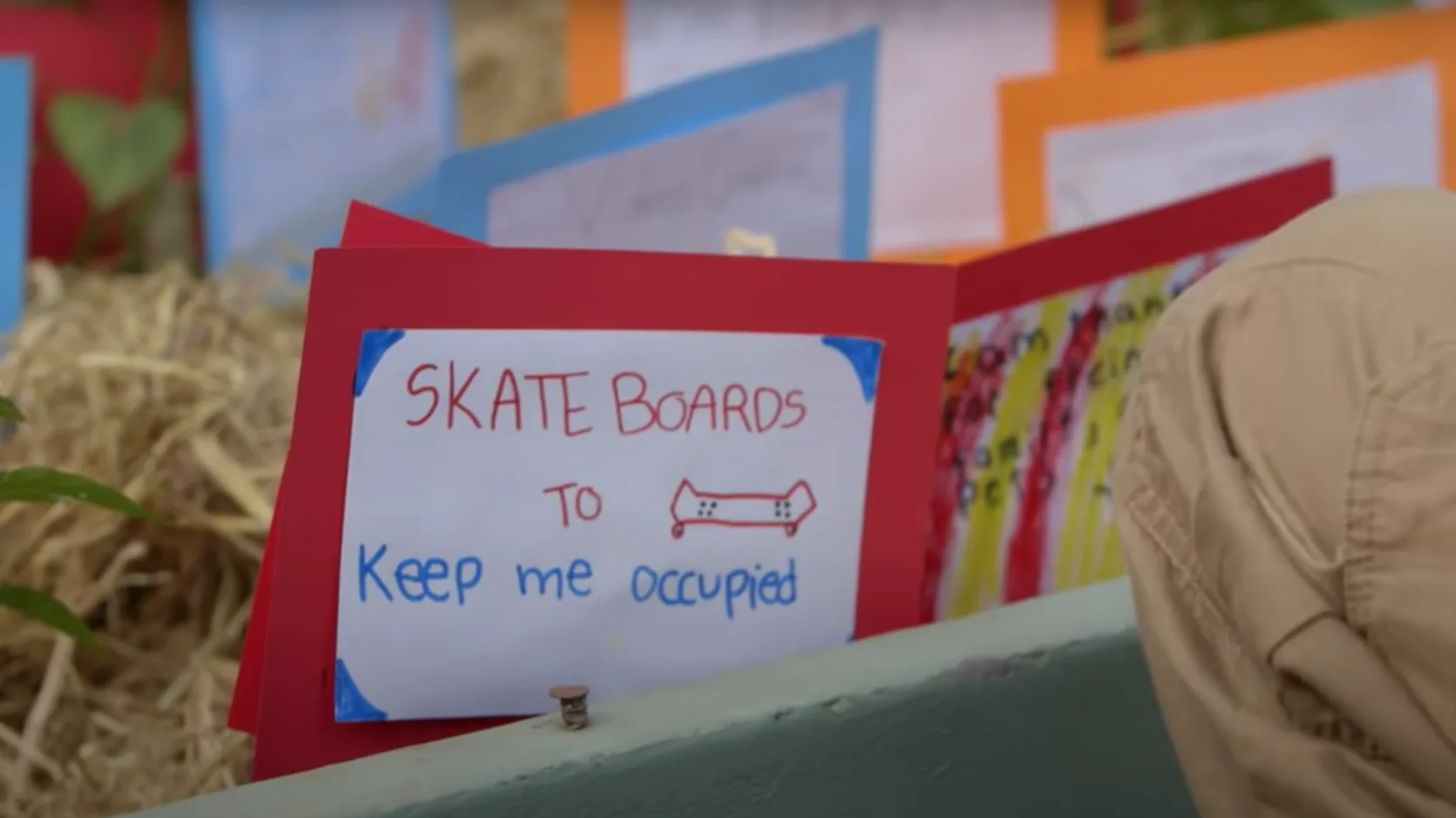 A handmade sign on red paper reads SKATE BOARDS TO keep me occupied with a drawing of a skateboard. Colorful papers and a persons knee are visible nearby.