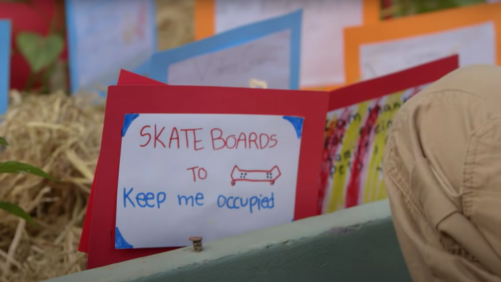 A handmade sign on red paper reads SKATE BOARDS TO keep me occupied with a drawing of a skateboard. Colorful papers and a persons knee are visible nearby.