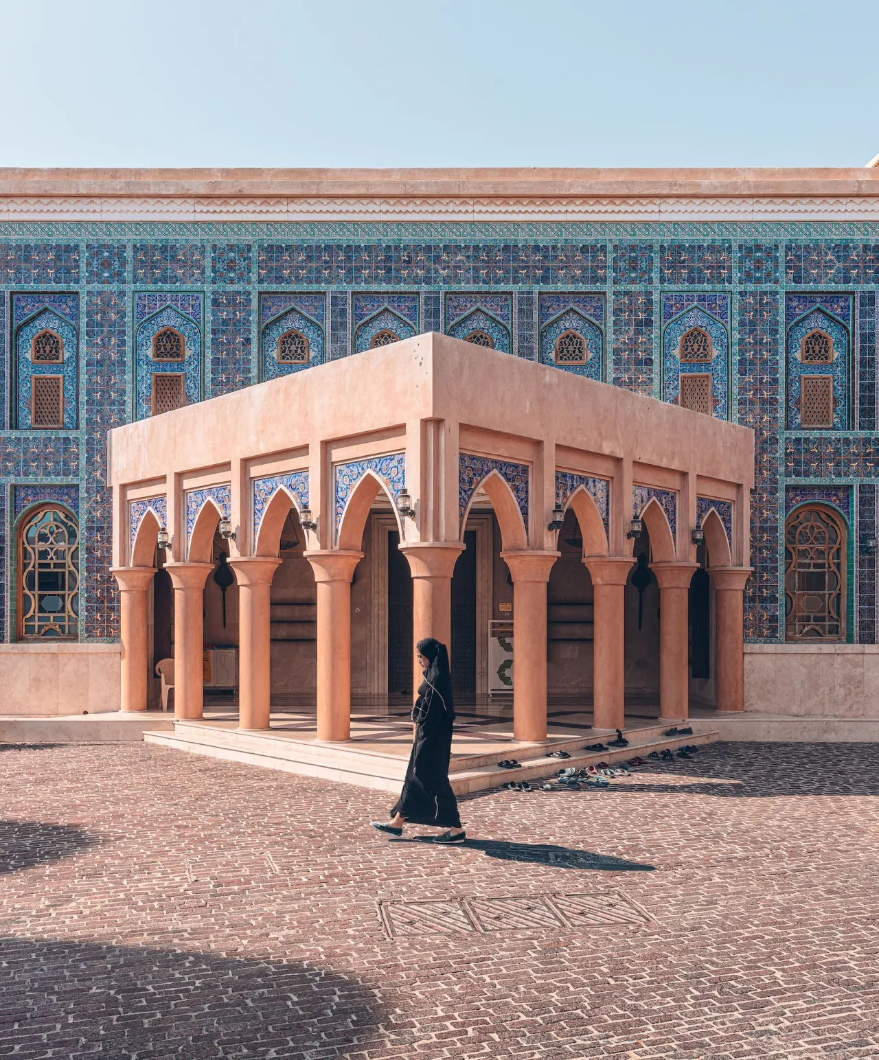 A person in black attire walks past a pavilion with arched columns in a courtyard. The background features a building with intricate blue mosaic patterns and arched windows under a clear sky.