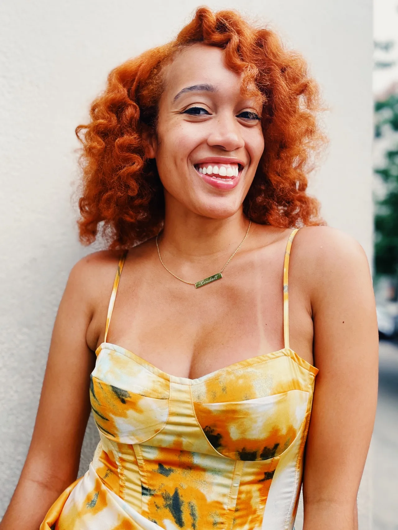 A smiling woman with curly, reddish hair wearing a yellow and white patterned dress and a necklace, standing outdoors against a light-colored wall.