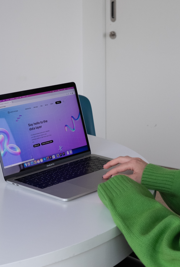 A person wearing a green sweater is using a laptop on a round white table. The laptop screen displays a website with purple and blue graphics and the text, Say hello to the data layer!.