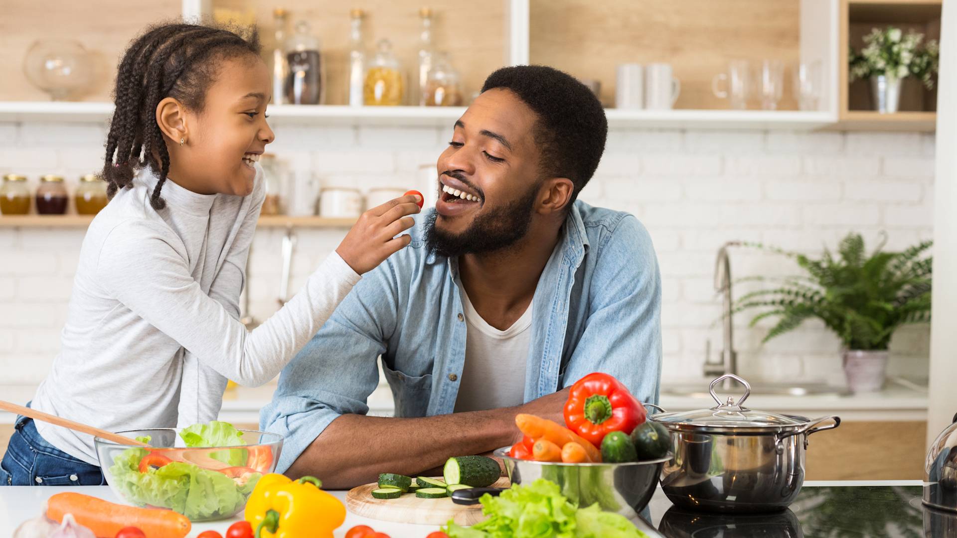 A young girl happily feeds a smiling man, likely her father, a slice of tomato while they prepare fresh vegetables together in a bright kitchen. Various colorful vegetables and a pot are on the counter.