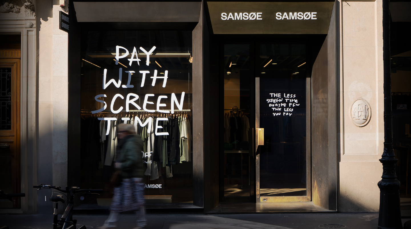 A clothing store with large windows displays the text PAY WITH SCREEN TIME in bold, white letters. The door shows the message The less screen time, the less you pay. A person walks by with a bicycle in front.