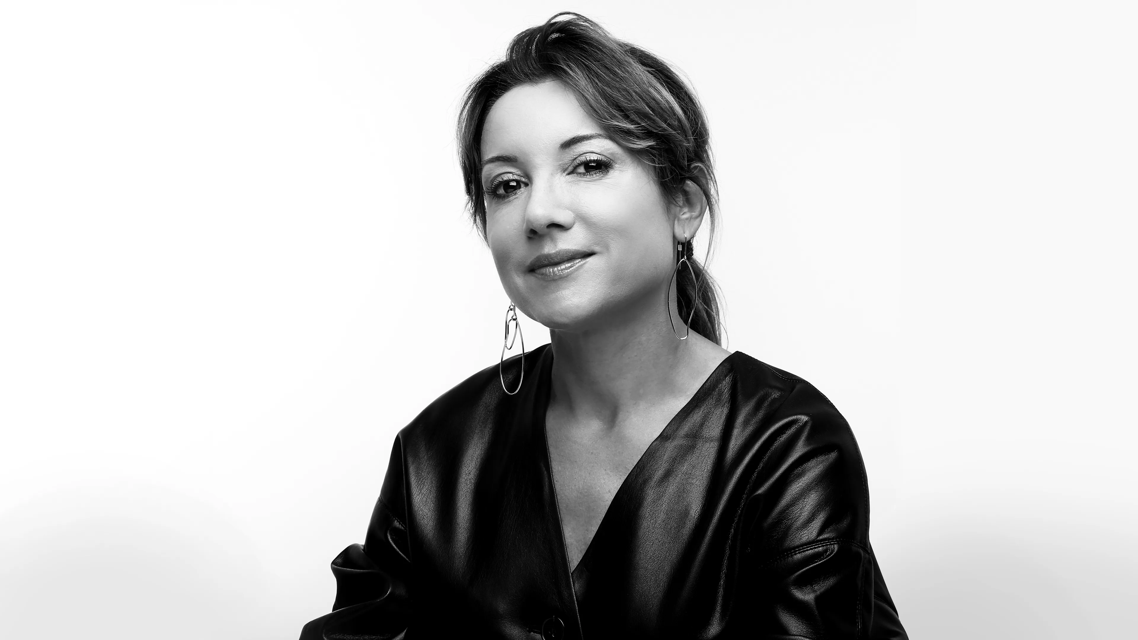Black and white portrait of a woman with medium-length hair pulled back, wearing a V-neck, leather-like top and large hoop earrings, looking at the camera with a slight smile against a plain background.