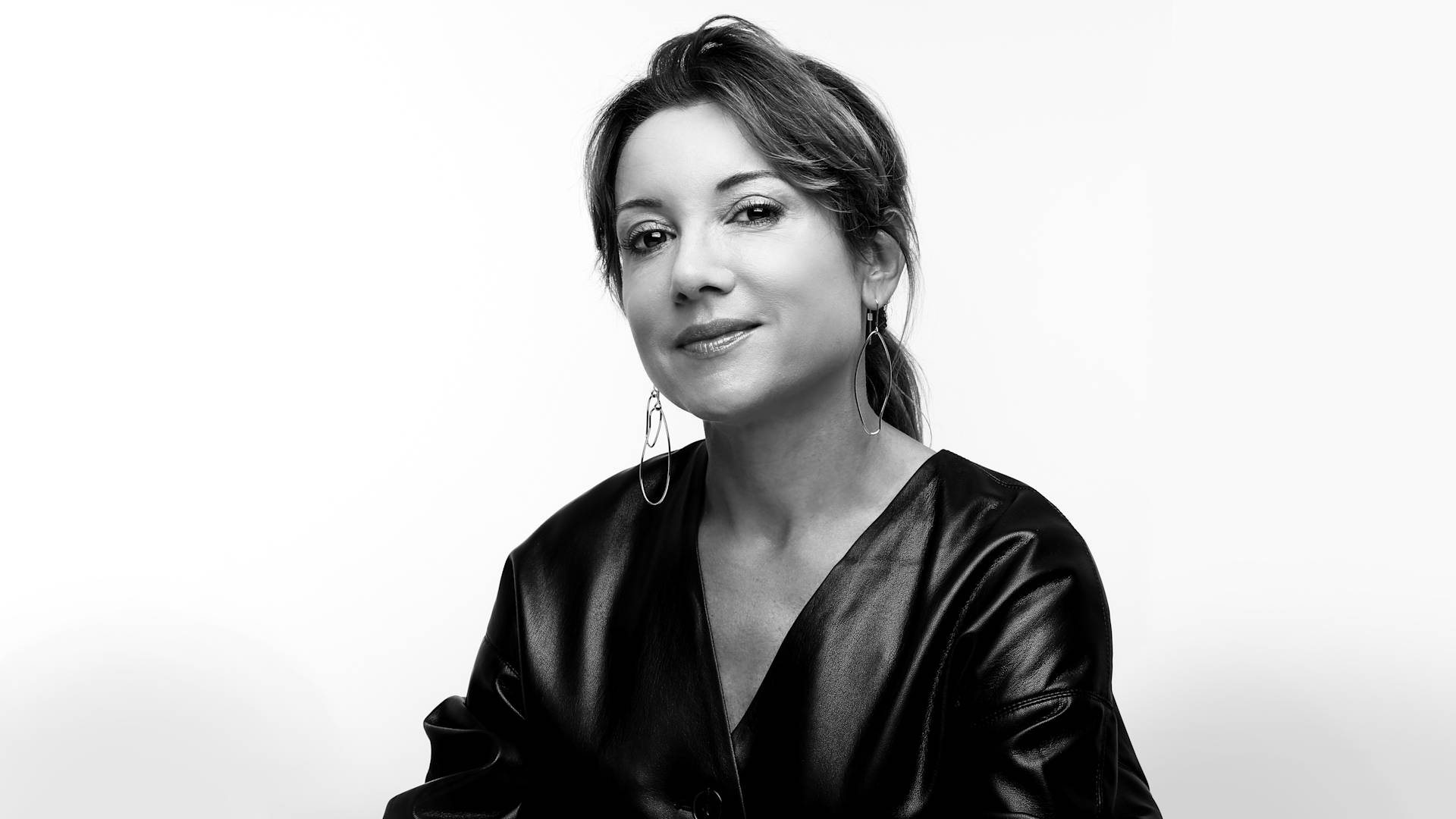 Black and white portrait of a woman with medium-length hair pulled back, wearing a V-neck, leather-like top and large hoop earrings, looking at the camera with a slight smile against a plain background.