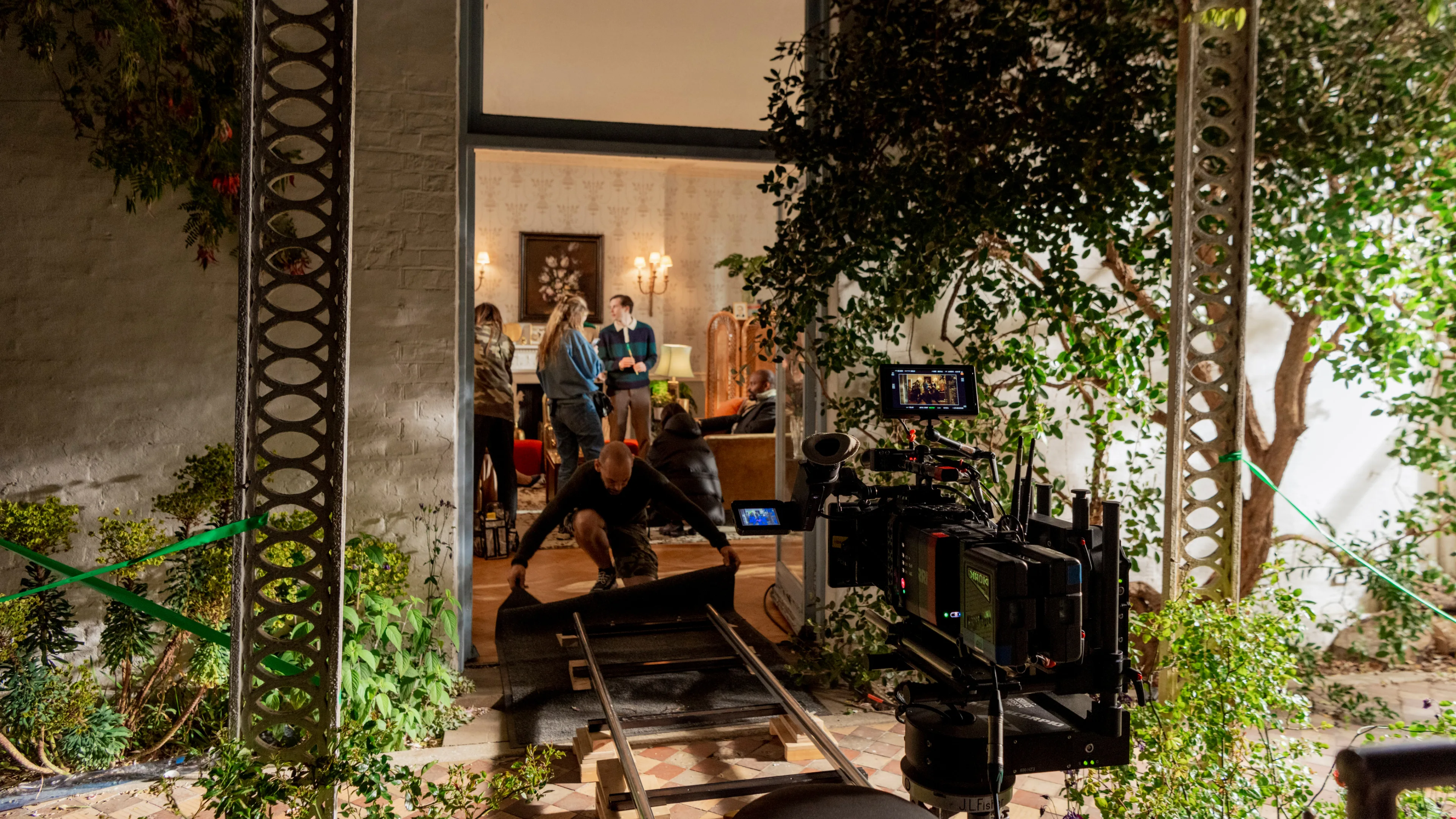 People set up a film scene inside a room as crew members adjust equipment and roll out a black mat on the floor. A camera on a track faces the open doorway, surrounded by plants and lighting gear.