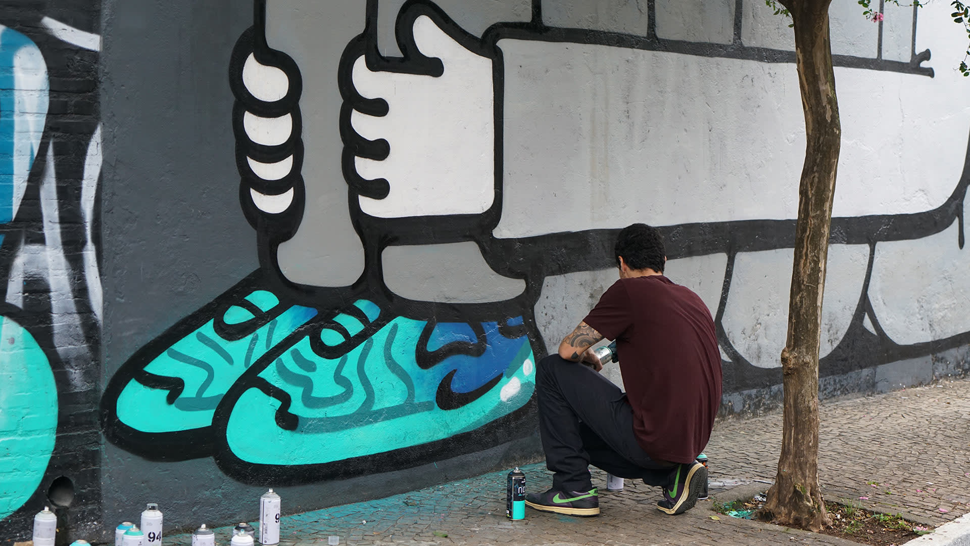 A person crouches on the sidewalk, spray painting a large mural on a wall. The mural features oversized turquoise shoes and bold black outlines. Several spray paint cans are scattered on the ground nearby.