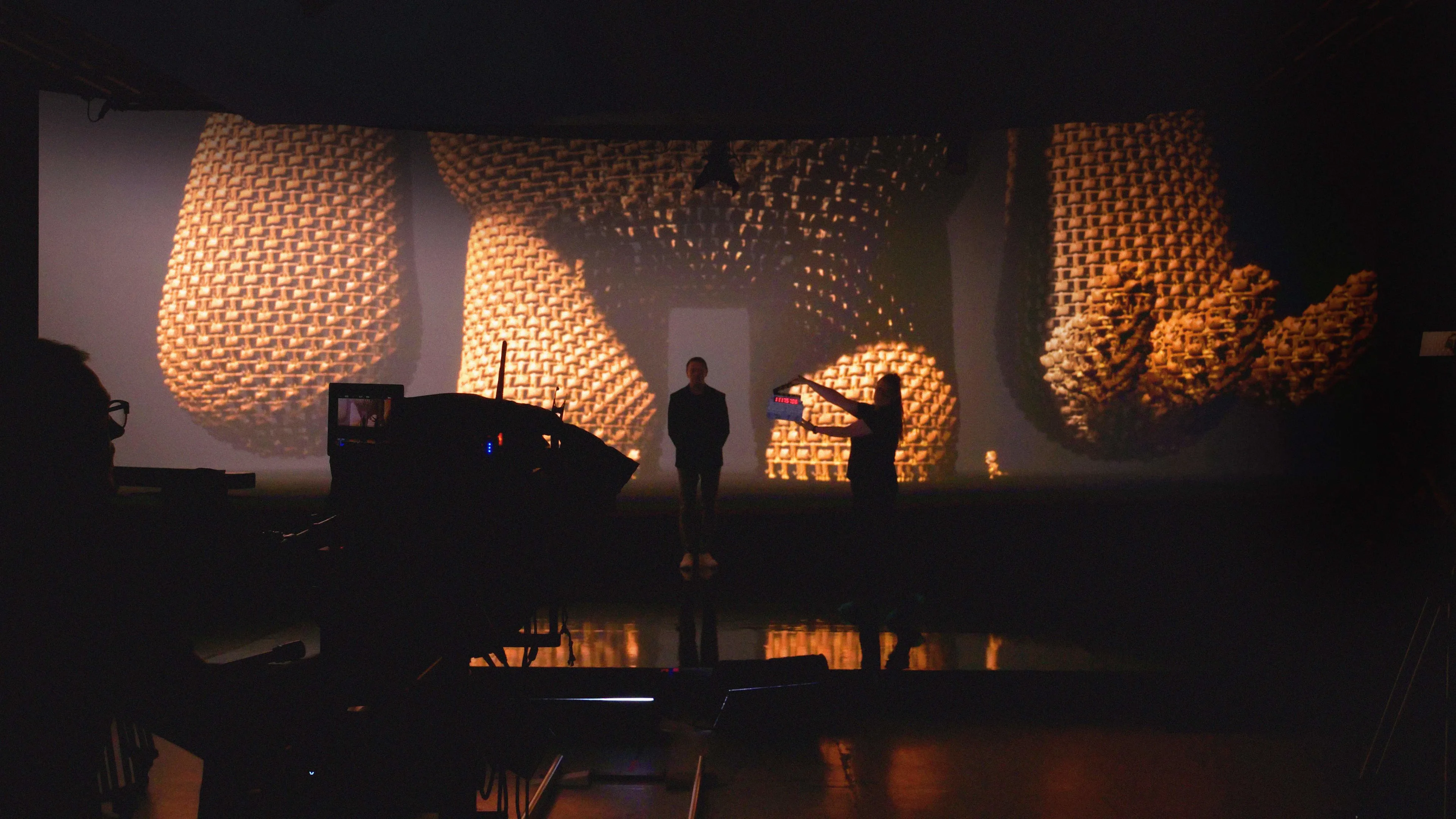 A person stands on a dimly lit stage with another person filming them. The background displays an enlarged, textured 3D object resembling woven material and large sculpted hands lit in warm tones.