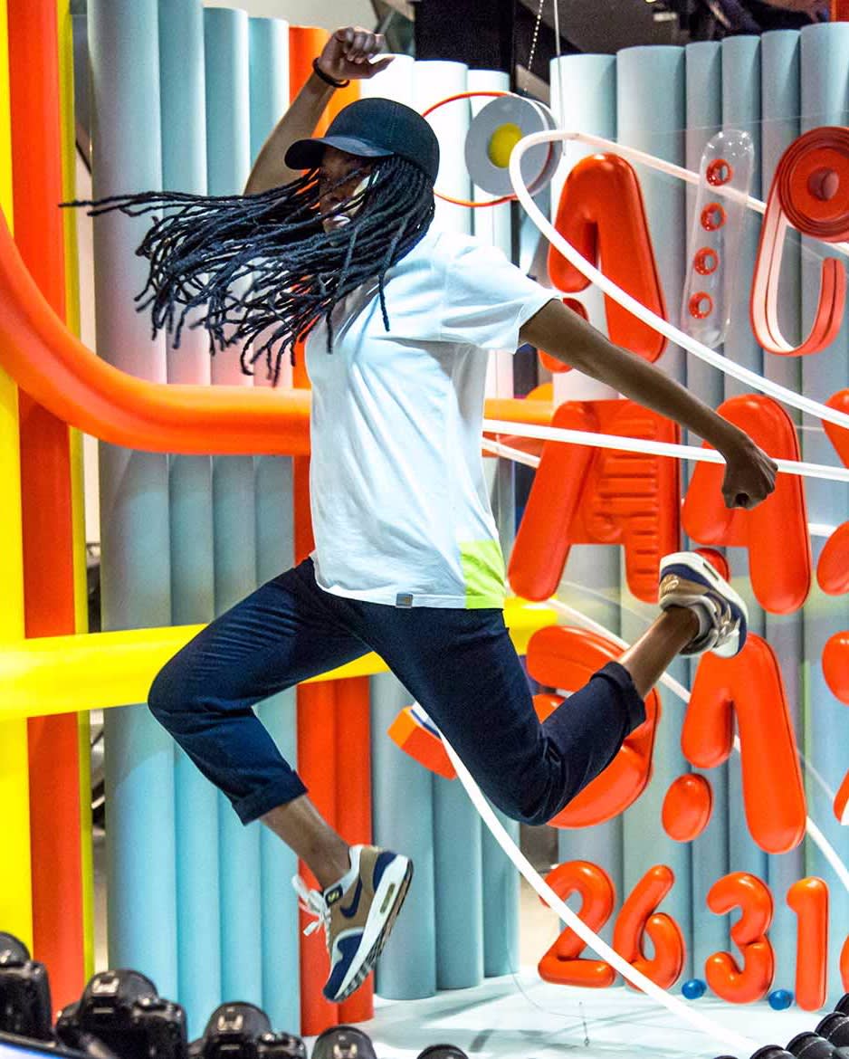 A person with long braided hair, wearing a white t-shirt, dark pants, sneakers, and a cap, jumps energetically in front of a colorful, abstract background with orange tubes and bold text.
