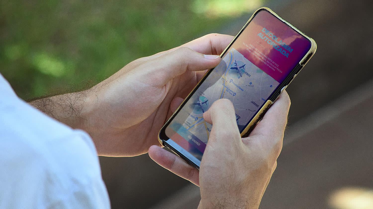 A person holds a smartphone displaying an airline app with a map and flight information on the screen, using their finger to interact with it. The background is blurred greenery and pavement.