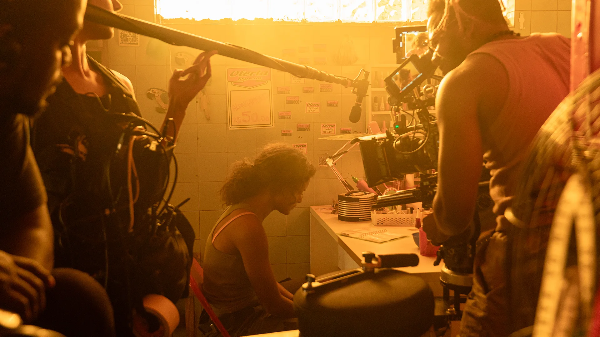A film crew shoots a scene of a person sitting at a small table in a warmly lit, cluttered room. The camera, microphones, and crew are visible, capturing the intimate moment.