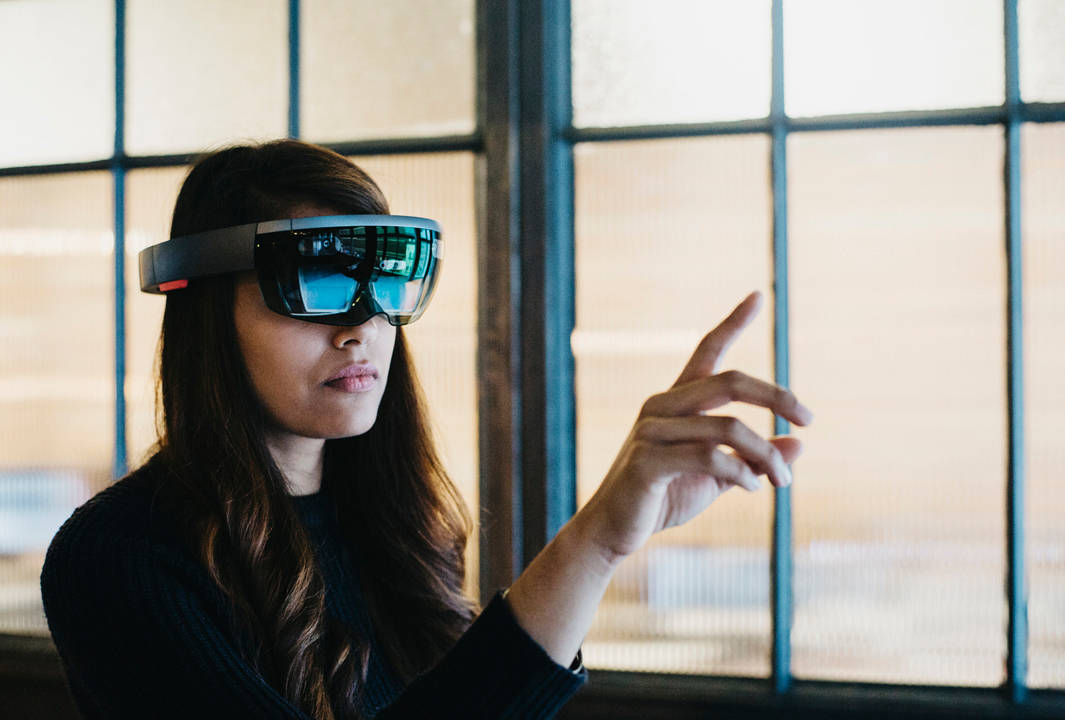 A woman wearing augmented reality glasses interacts with a virtual interface, pointing her finger in the air while standing indoors near large windows.