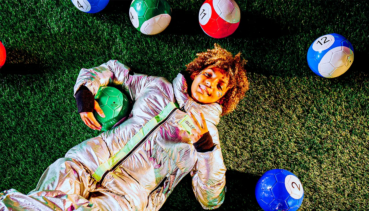 A person in a shiny silver jacket lies on artificial grass, holding a green soccer ball. Around them are several colorful soccer balls, some with numbers, arranged on the turf.