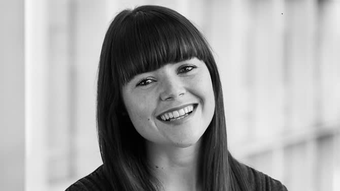 A woman with straight dark hair and bangs smiles at the camera in a black and white photo. The background is softly blurred, giving a professional and friendly impression.