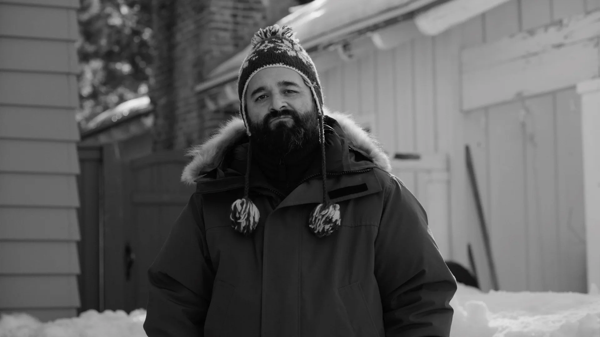 A man with a beard stands outside in the snow, wearing a winter jacket with a fur-lined hood and a patterned knit hat with tassels. The scene is in black and white, and snow surrounds him.
