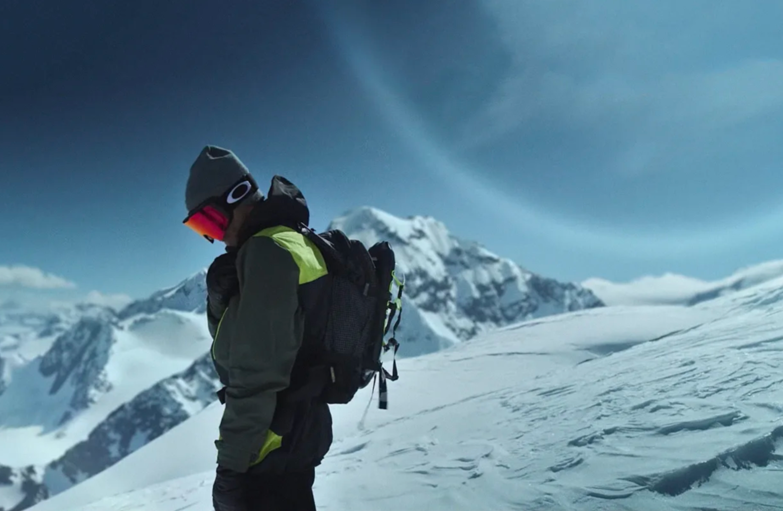 A person wearing winter gear and a backpack stands on a snowy mountain slope with snow-covered peaks and a cloudy, blue sky in the background.