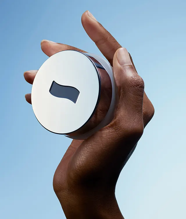 A hand holding a round, white container with a metallic rim and a wavy, dark window on the lid, set against a light blue background.
