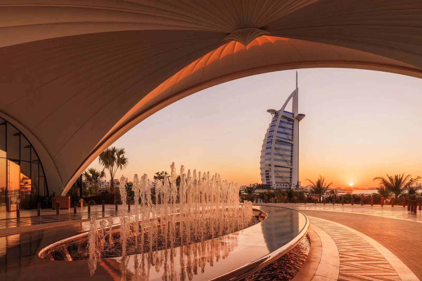 Curved modern architecture with a fountain in the foreground, palm trees, and the Burj Al Arab hotel in the background at sunset in Dubai, United Arab Emirates.