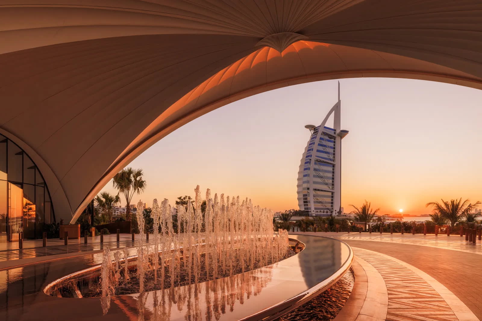 Curved modern architecture with a fountain in the foreground, palm trees, and the Burj Al Arab hotel in the background at sunset in Dubai, United Arab Emirates.