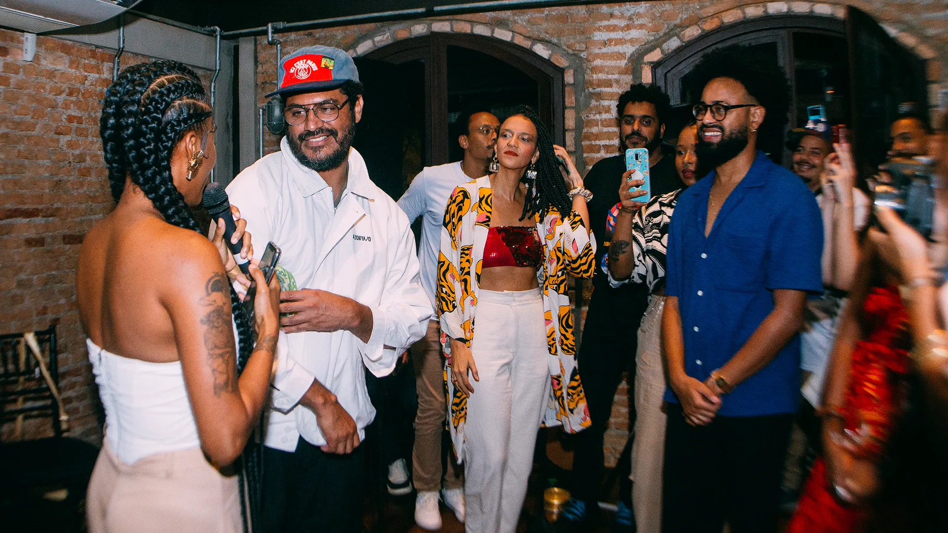 A woman with braided hair speaks into a microphone to a smiling man in a white jacket and red cap at a lively indoor gathering, while others watch, smile, and take photos with their phones.