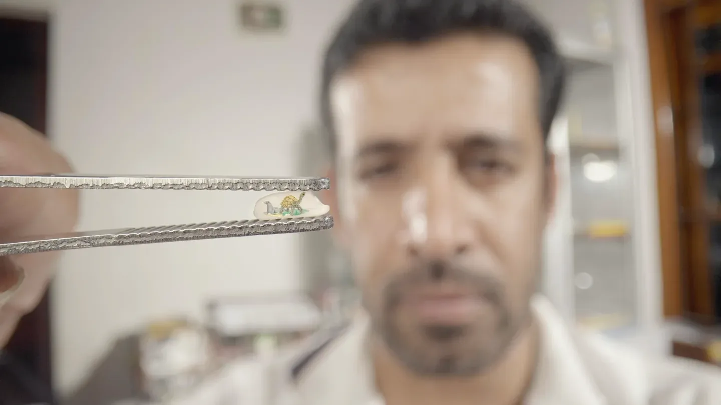 A man holds a pair of tweezers displaying a tiny, intricate sculpture of a chameleon, with the mans face blurred in the background.