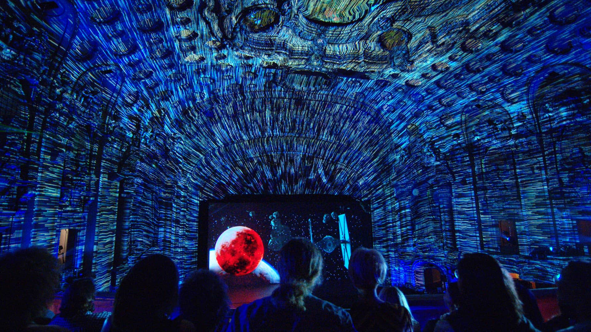 A group of people watch an immersive digital projection on a domed ceiling, featuring swirling blue and yellow patterns, a red planet, and cosmic imagery, creating a vibrant, space-themed environment.