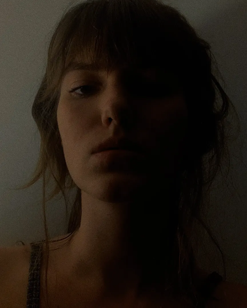 A person with long hair and bangs is lit from the side, casting much of their face in shadow. They have a neutral expression and are wearing a textured tank top. The overall lighting is dim and moody.